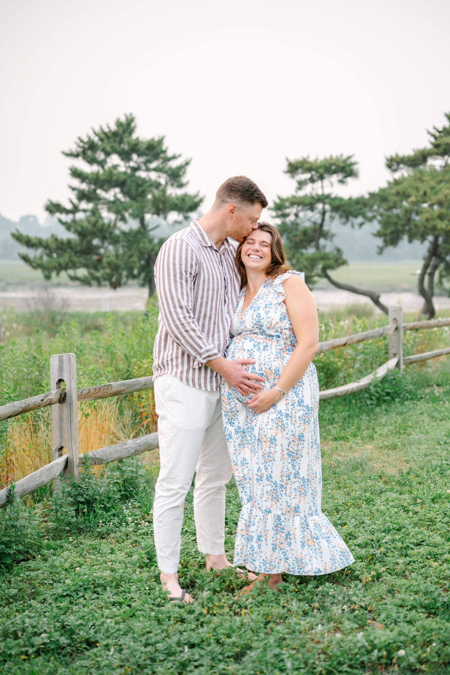 Couple embracing outdoors, pregnant woman in floral dress, man in striped shirt, rustic fence and green trees background.