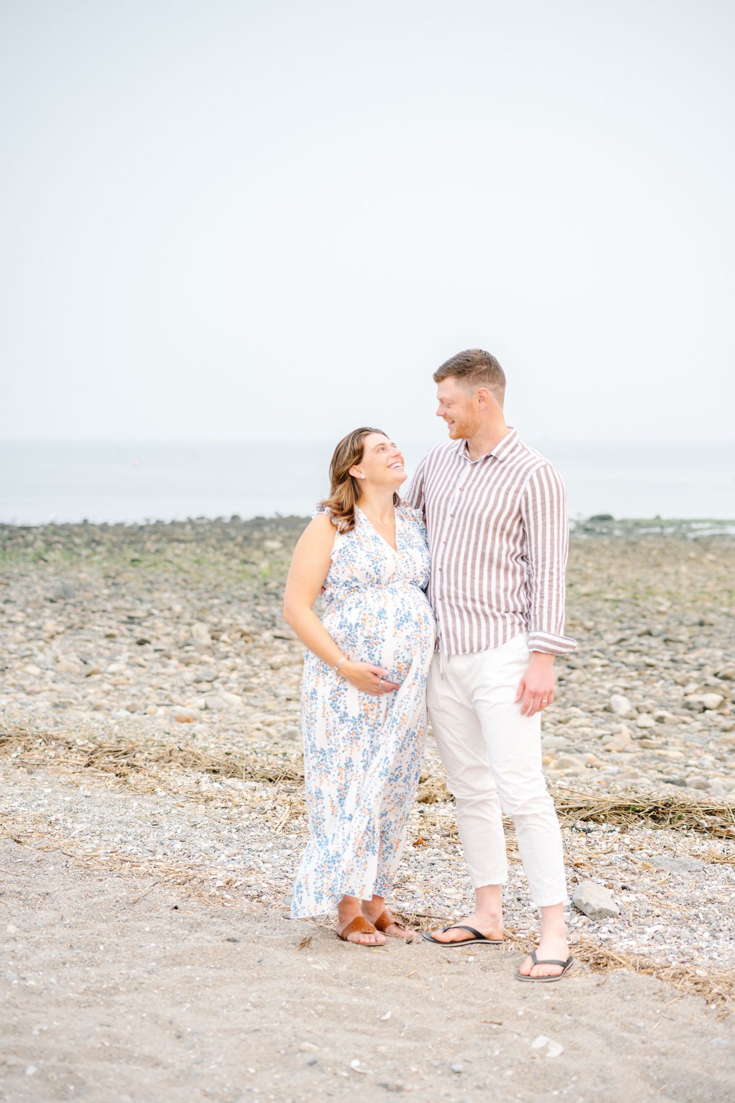 Pregnant couple smiling at each other on a rocky beach, woman in floral dress, man in striped shirt and white pants.