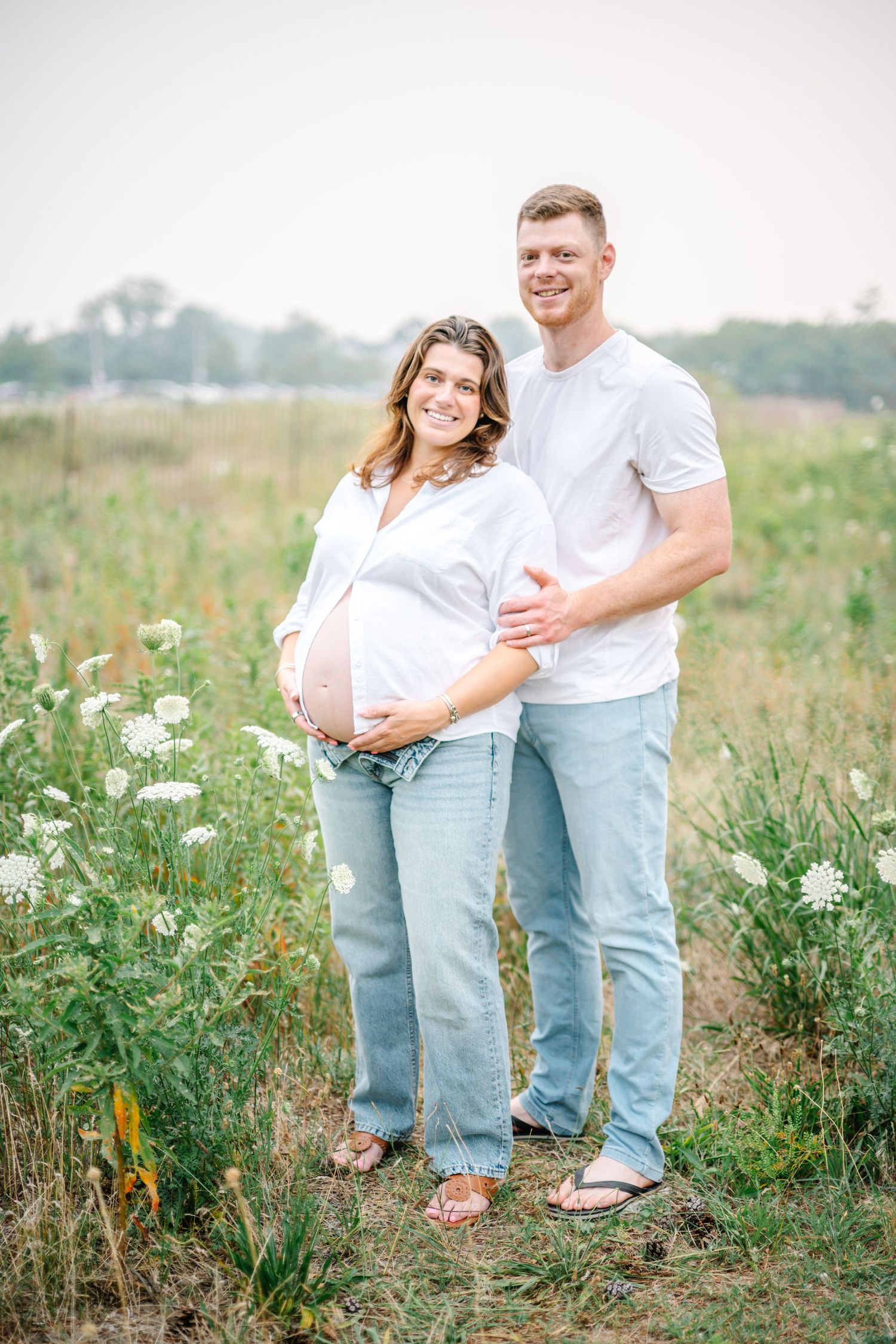 Smiling pregnant couple in white tops and jeans posing in a wildflower field during a maternity photo session.