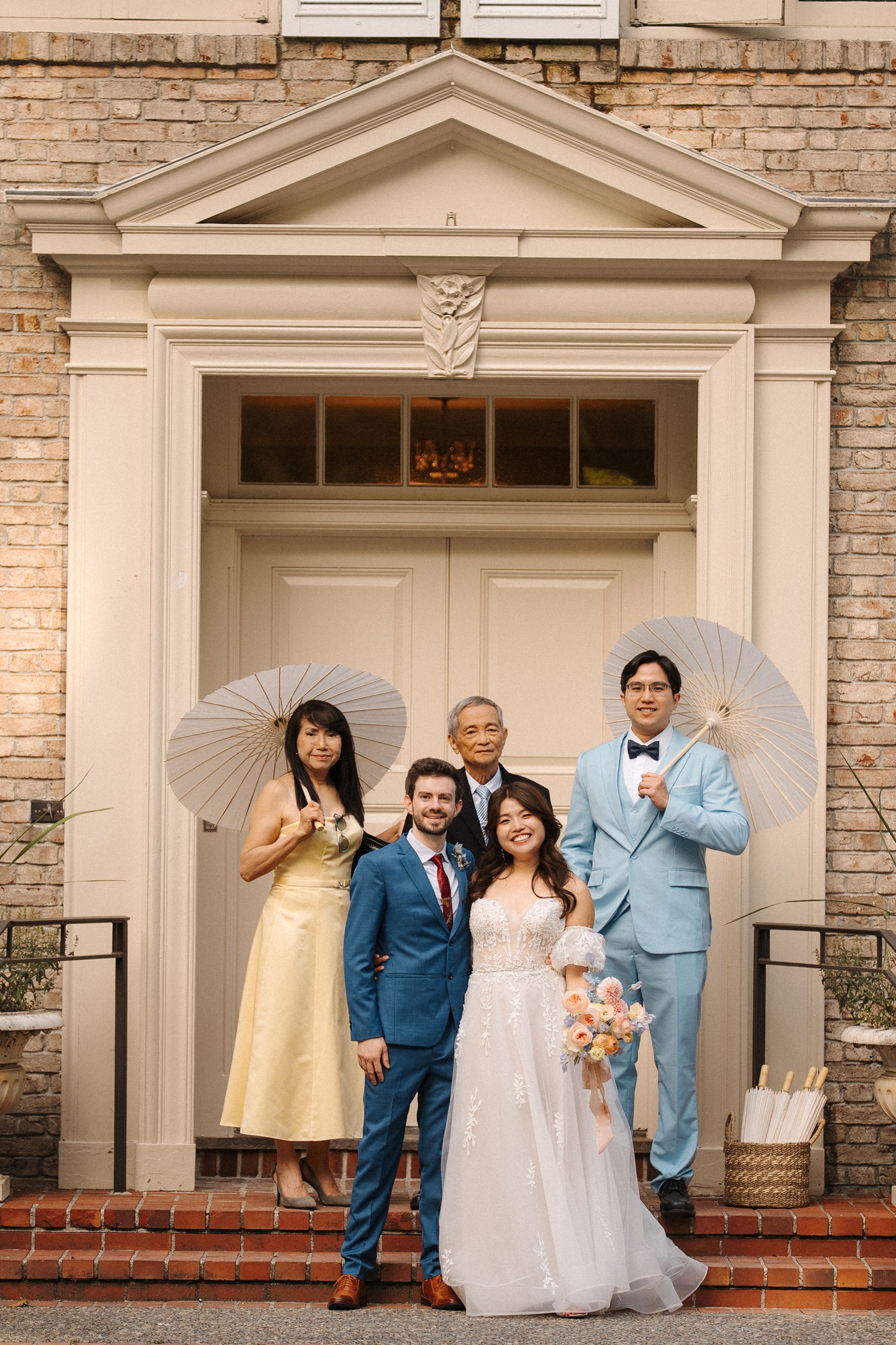 Wedding party poses playfully in front of brick building with white columns and doors during outdoor wedding photoshoot.