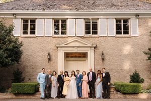 A wedding party arranged in a line poses for formal photos outside a grand stone building with white shuttered windows.