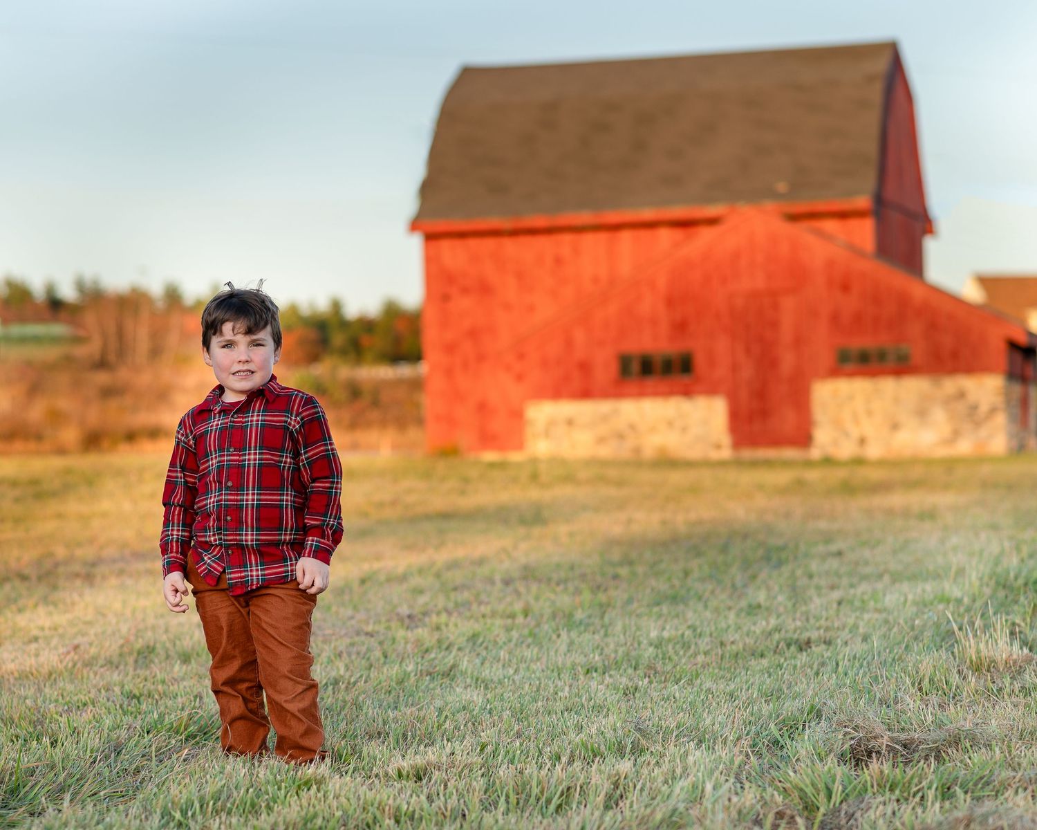 A child in a plaid shirt and brown pants stands in front of red barns in a rural autumn landscape.