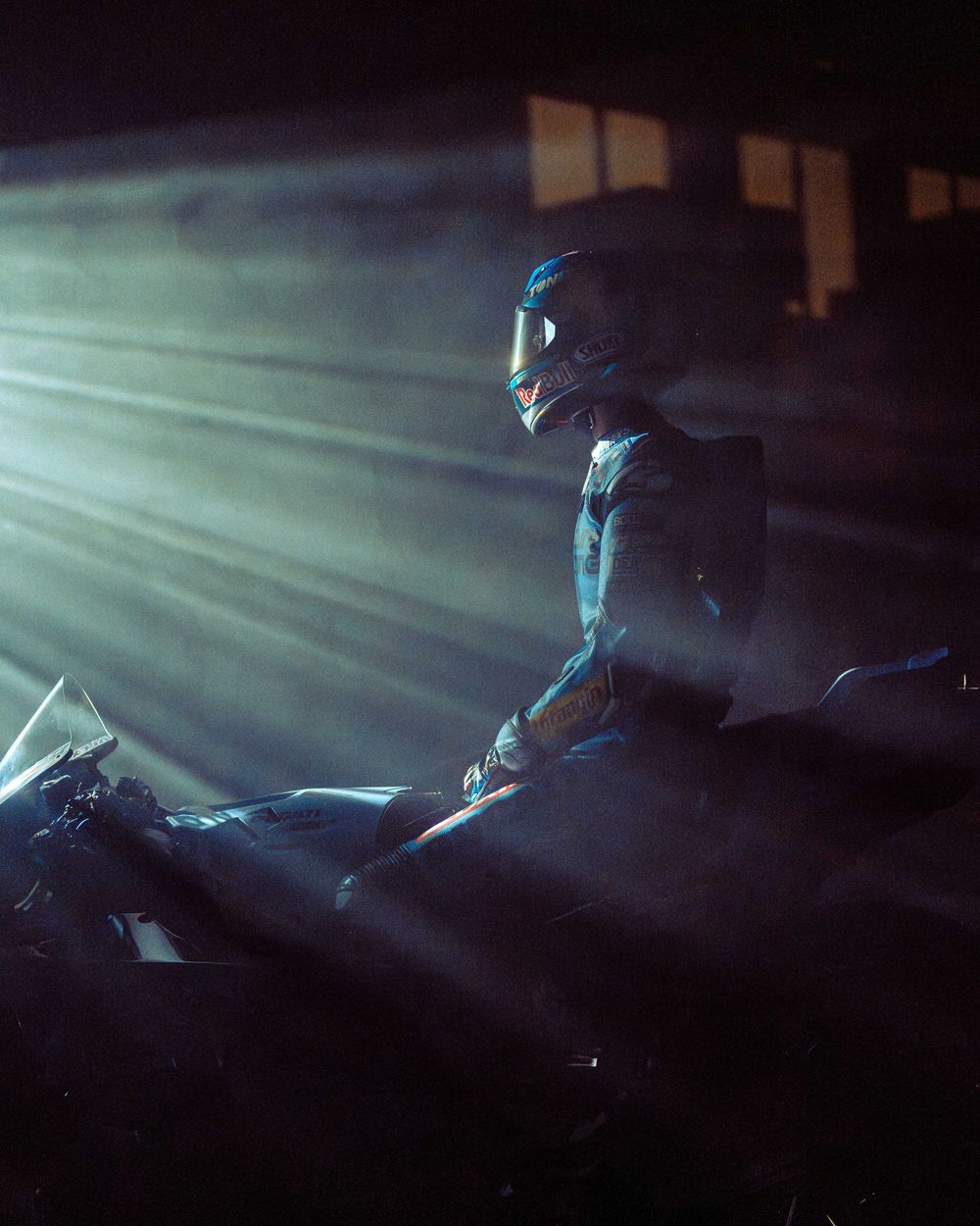 A person in a motorcycle helmet sits on a bike in dramatic lighting with beams of light cutting through fog.