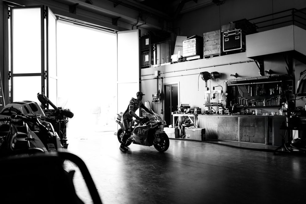 Black and white interior of a motorcycle repair shop with bikes and equipment visible in natural window light.