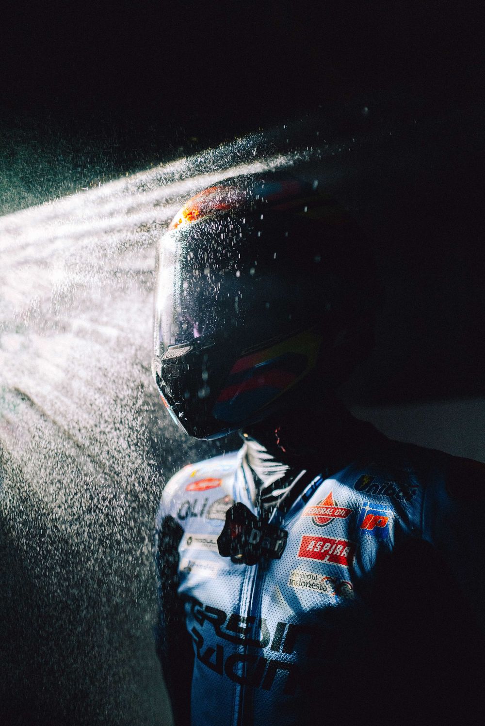 Dramatic high-contrast photo of a person in racing gear with water droplets spraying in dramatic lighting.