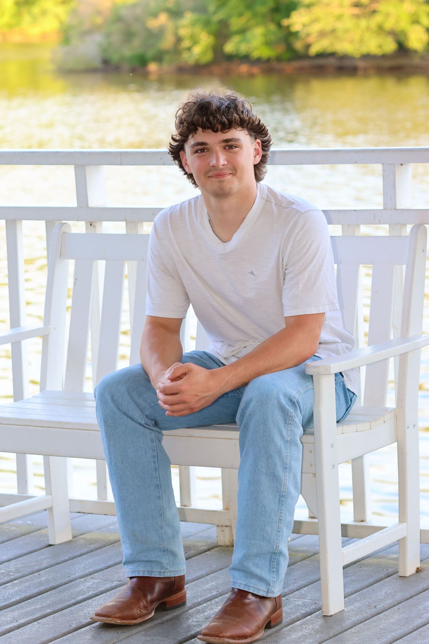 High school senior guy wearing a white t shirt and jeans sitting on a bench with a lake behind him.