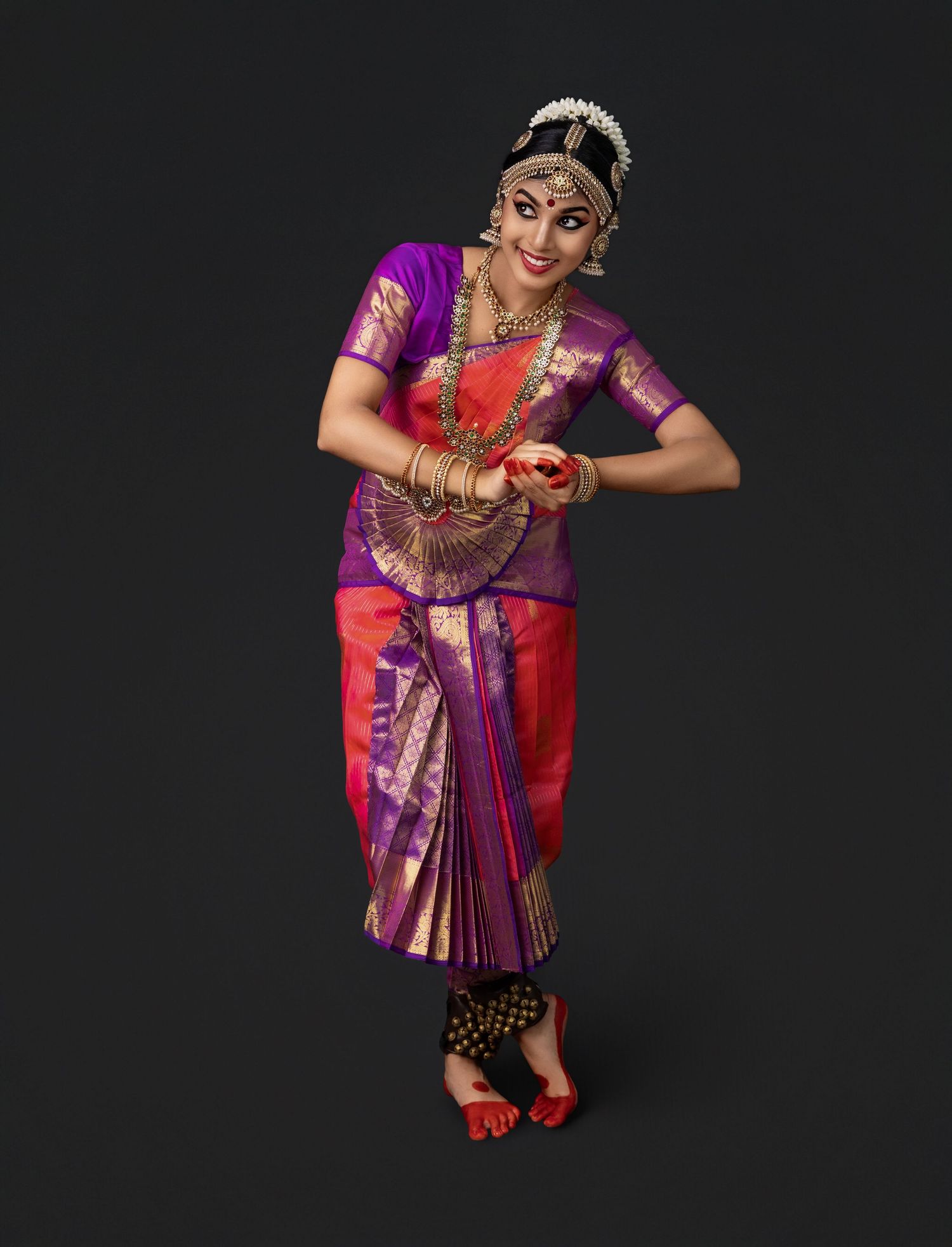 A dancer performs a traditional Bharatanatyam dance pose wearing a purple and red silk sari with ornate jewelry and accessories.