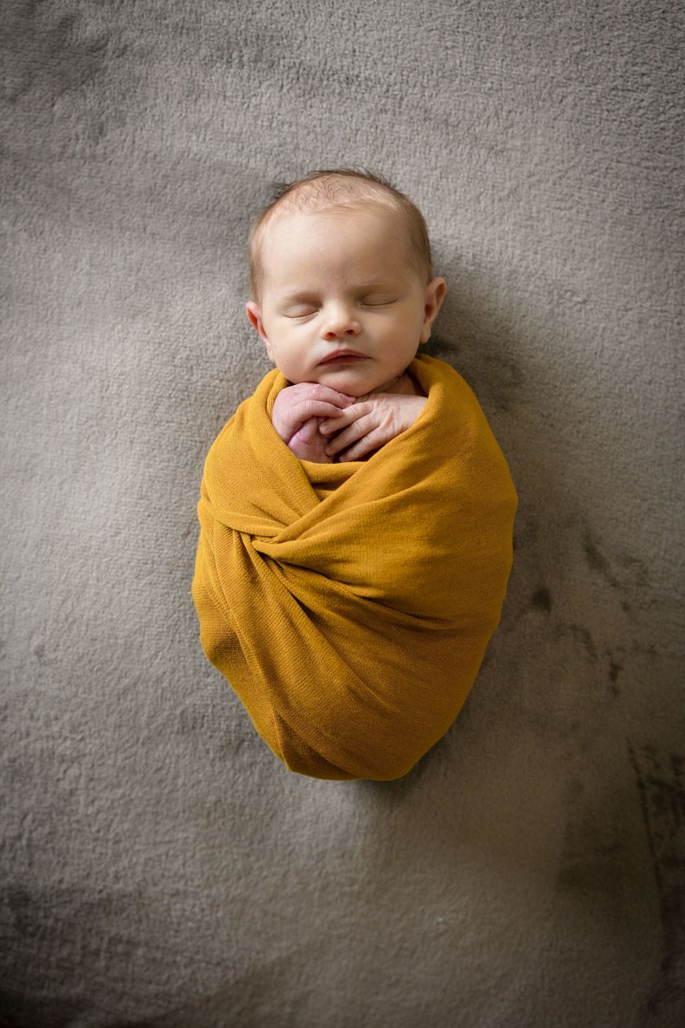 Sleeping newborn swaddled in mustard yellow wrap against grey backdrop.