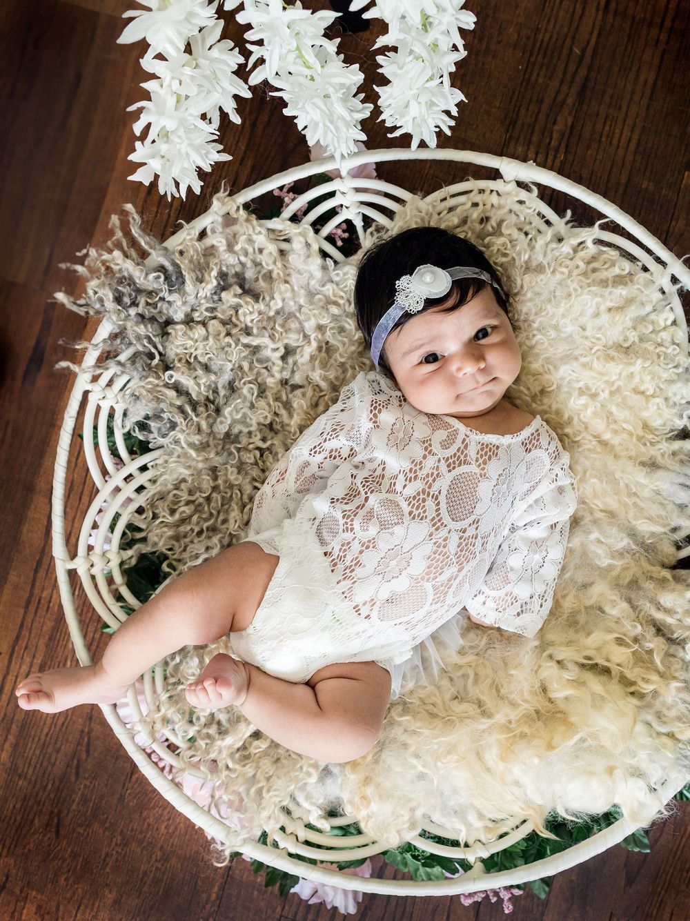 Newborn in lace dress lying in white decorative basket with flowers.