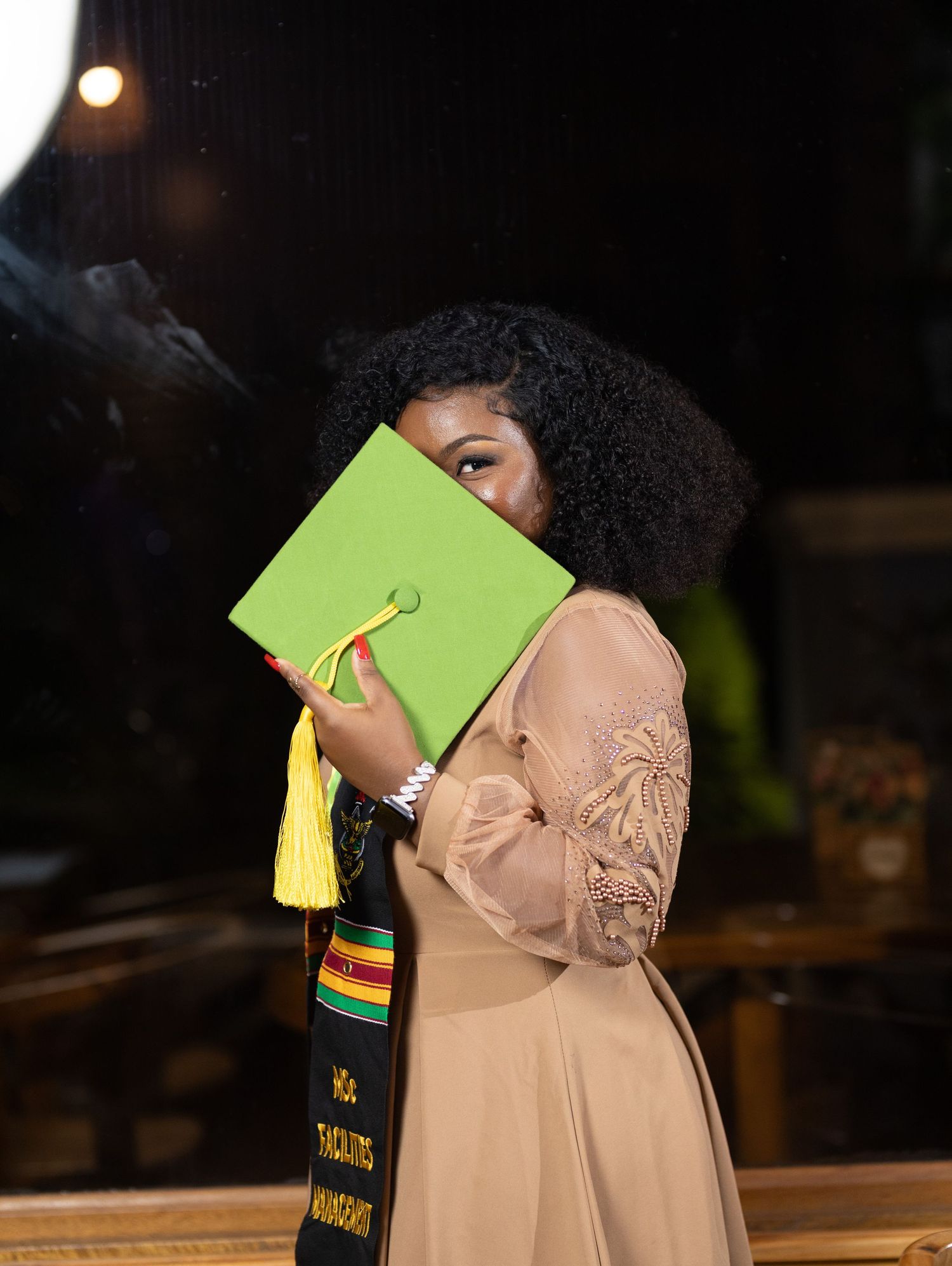 Graduate wearing academic regalia poses against wooden railing in modern interior space with dramatic lighting.