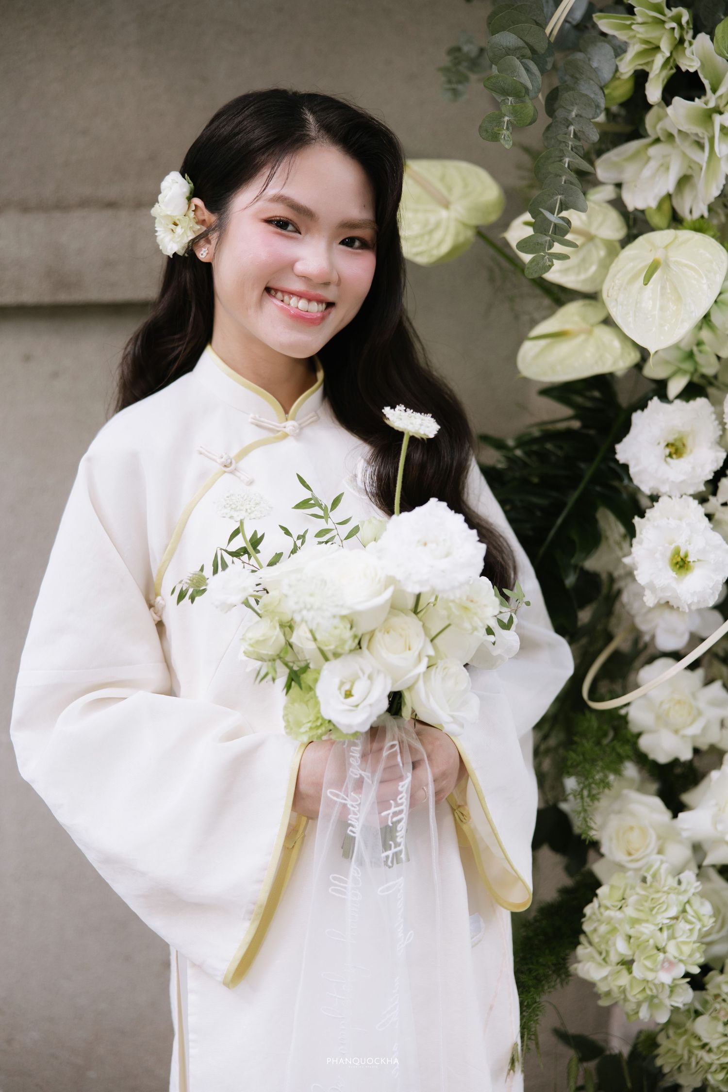 Smiling bride in white traditional Vietnamese ao dai holding white flower bouquet with floral backdrop.