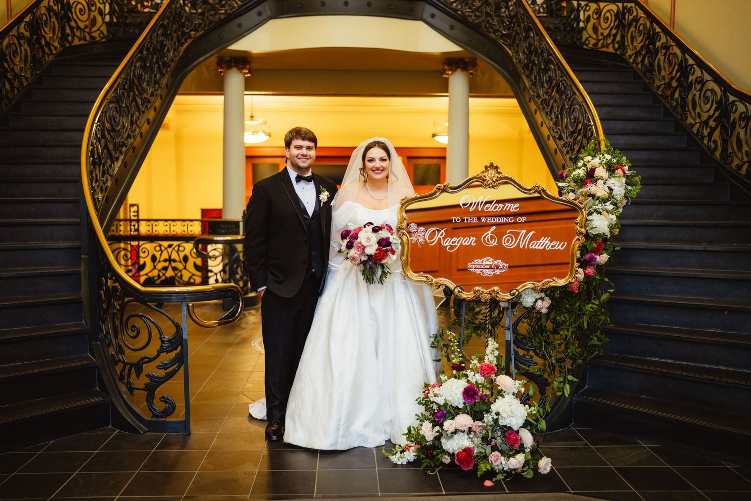 Couple stands at the bottom of an elegant staircase with ornate wrought iron railings and warm yellow lighting.