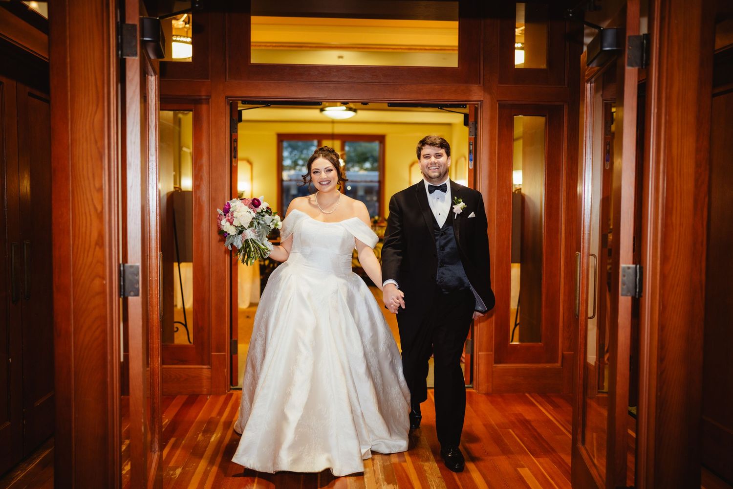 A bride and groom walk down an elegant wood-paneled hallway after their wedding ceremony.