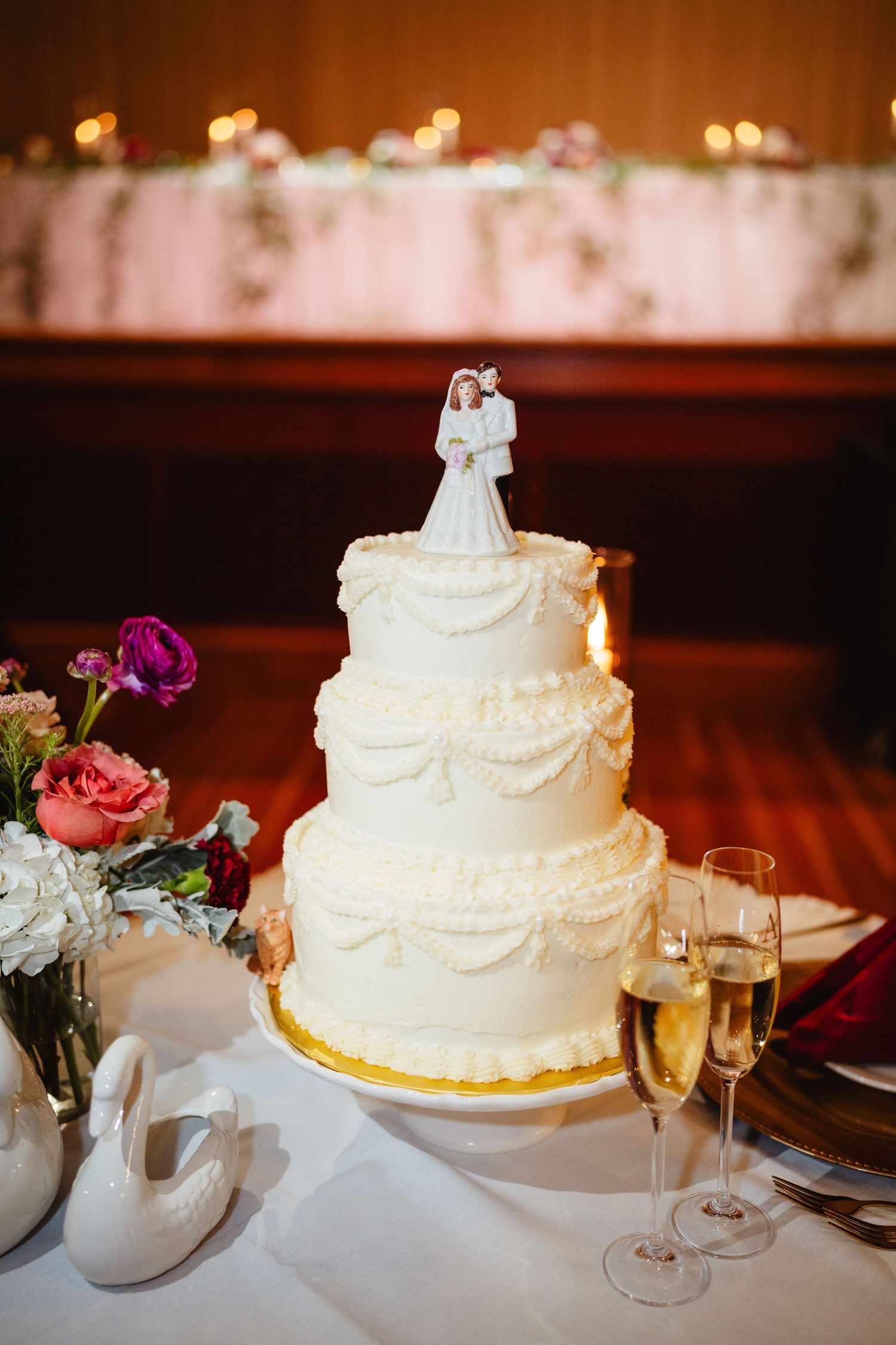 Traditional white wedding cake with delicate piped details and swags sits on reception table with champagne glasses.