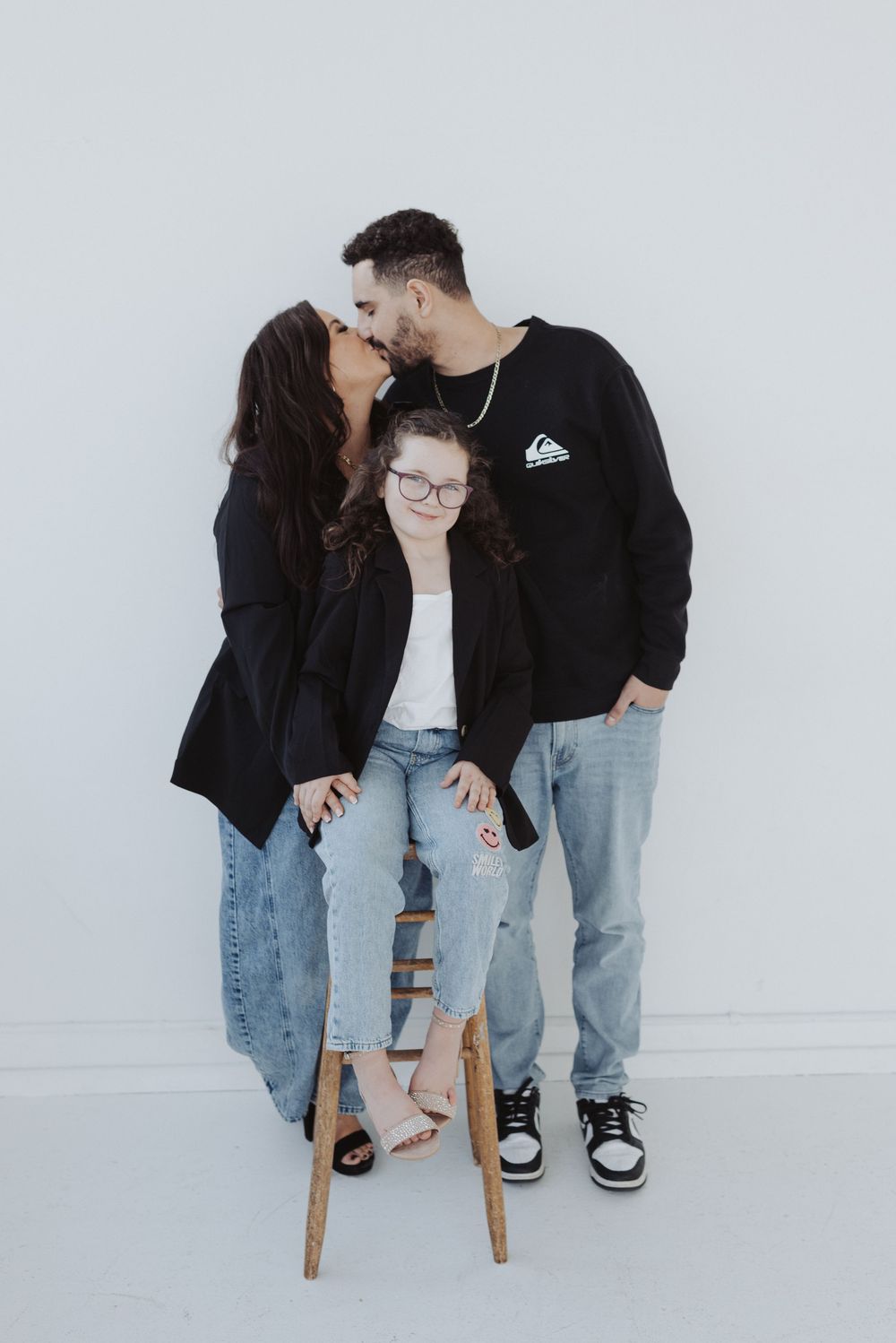 Couple in matching black tops and jeans pose together against white wall in minimalist photo series.