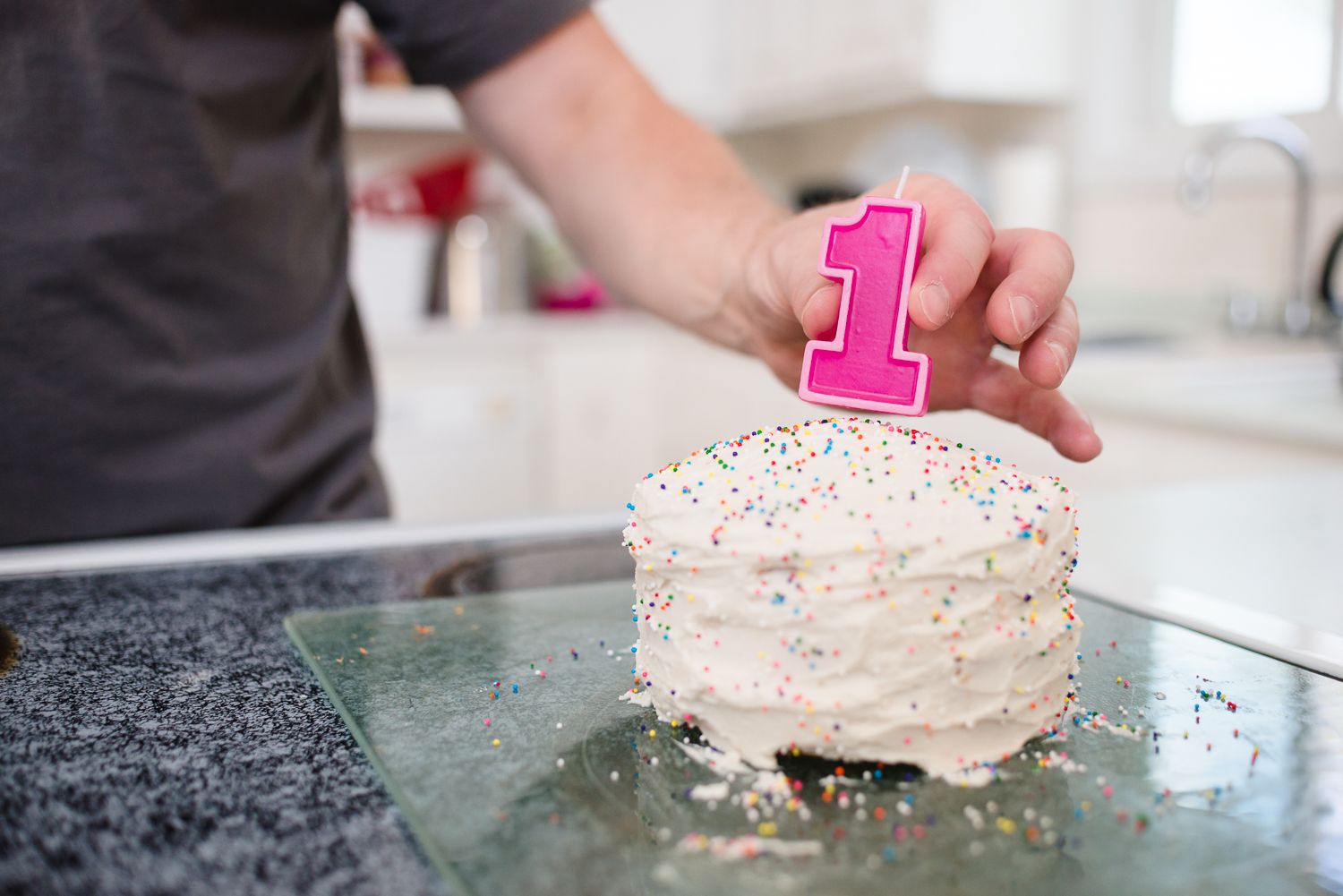 A plate with pink frosted birthday cake slice being decorated with colorful sprinkles and one candle.