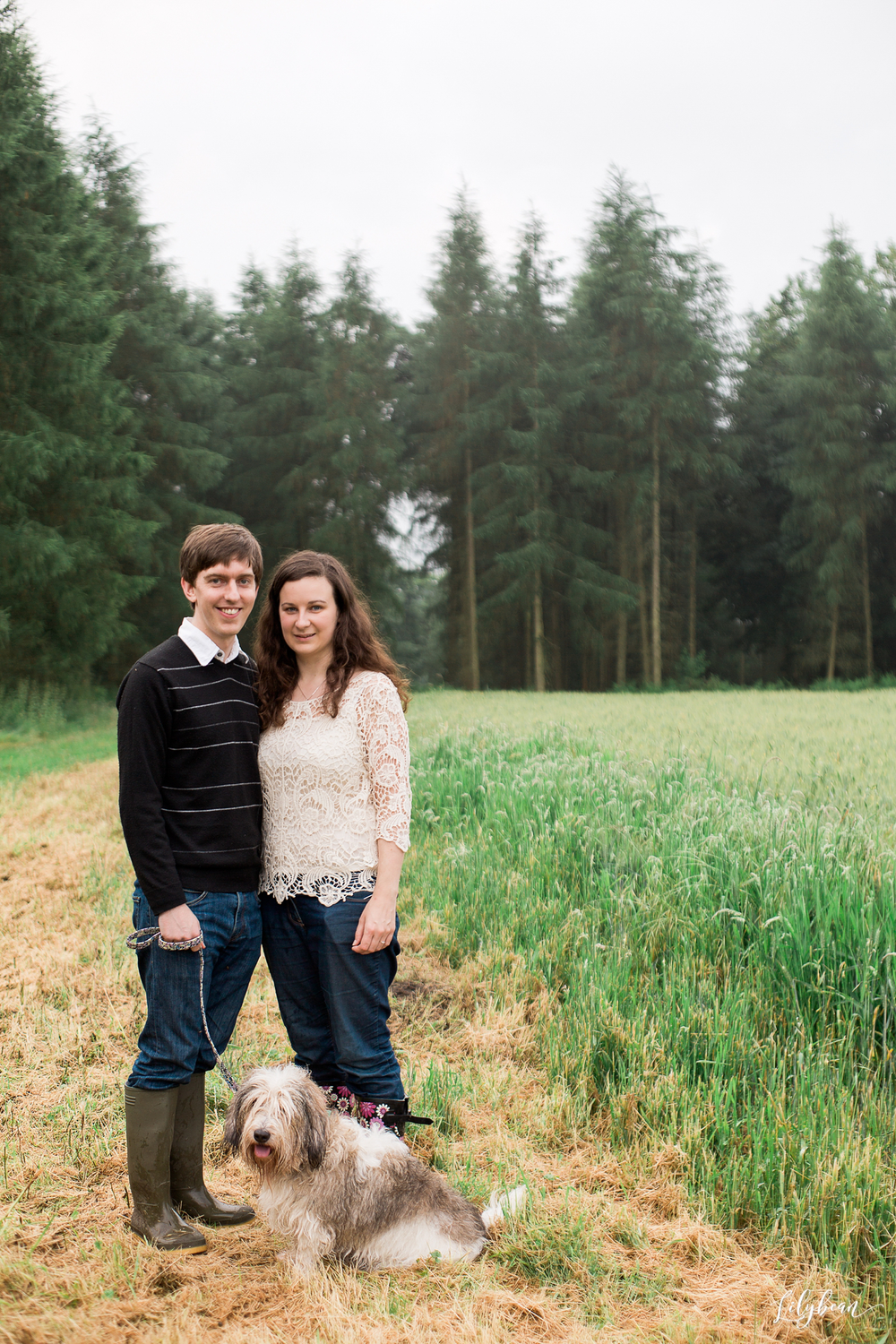 Couple poses with their shaggy dog in a green field with pine trees in the background.