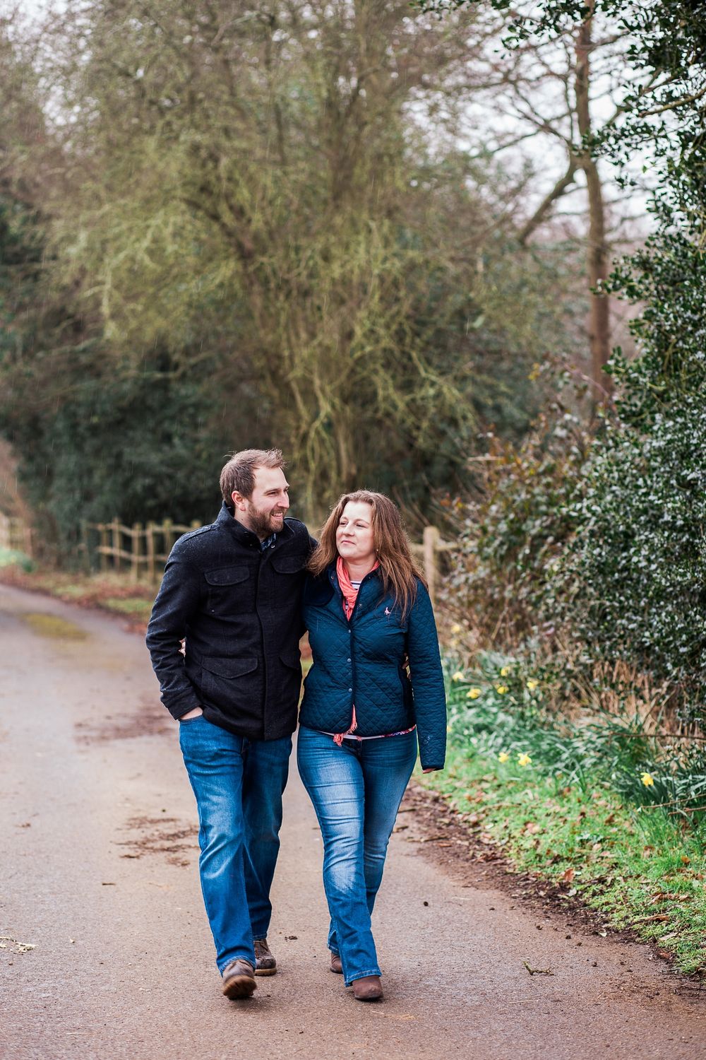 Couple walks together laughing on a tree-lined path wearing casual denim.