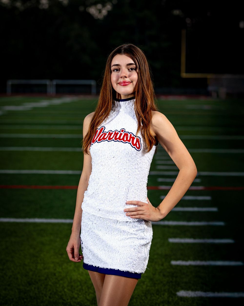 Cheerleader wearing sparkly white uniform stands on football field during golden hour.