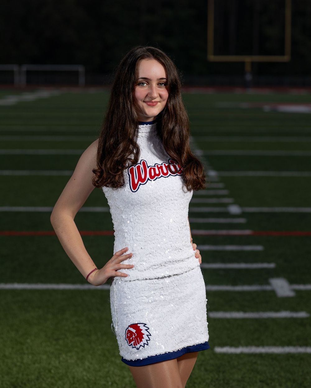 High school cheerleader in a white uniform poses on a football field with yard lines visible in the background.