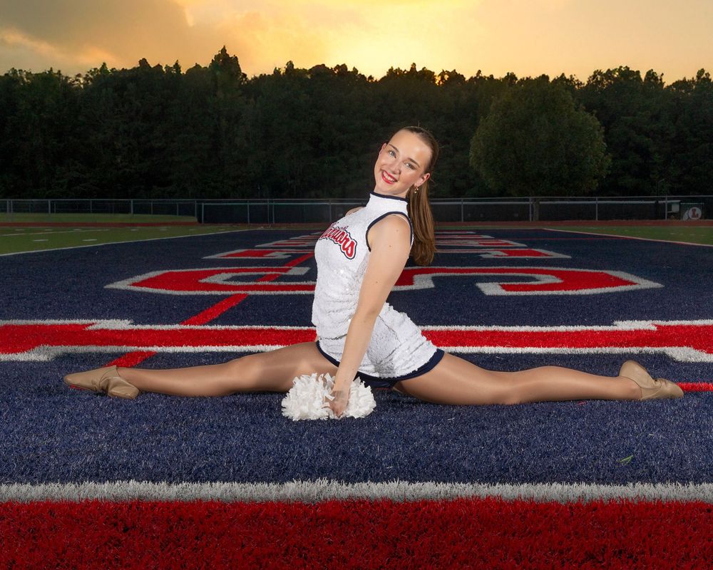 Cheerleader demonstrates flexibility with splits on red and blue football field at sunset.