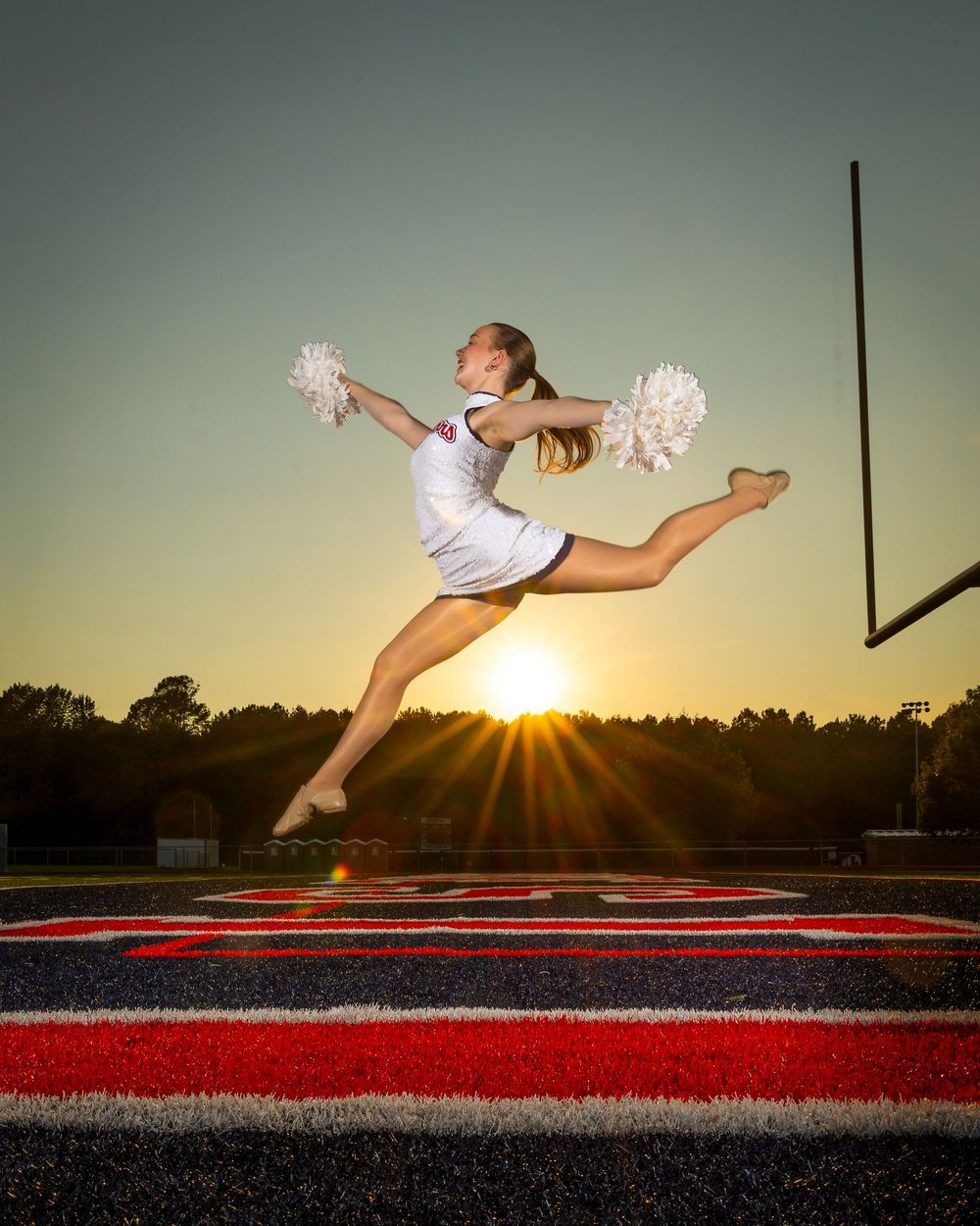 Cheerleader performs dramatic jump split against sunset sky on football field.