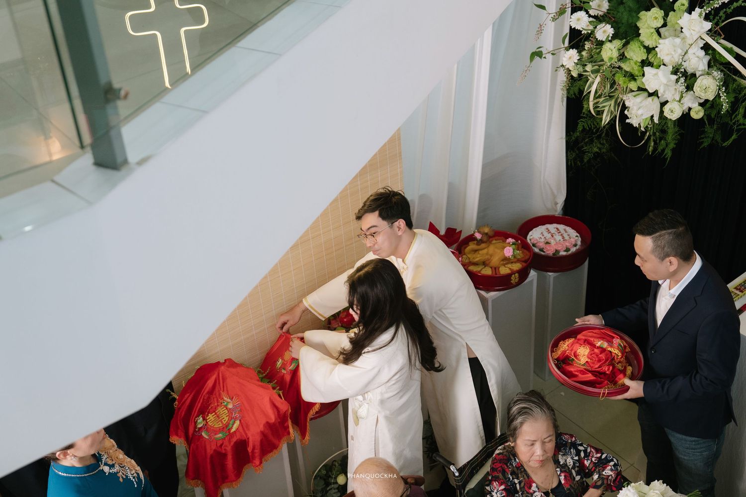 Overhead view of Vietnamese engagement ceremony with couple in white ao dai receiving red lacquer gift boxes.