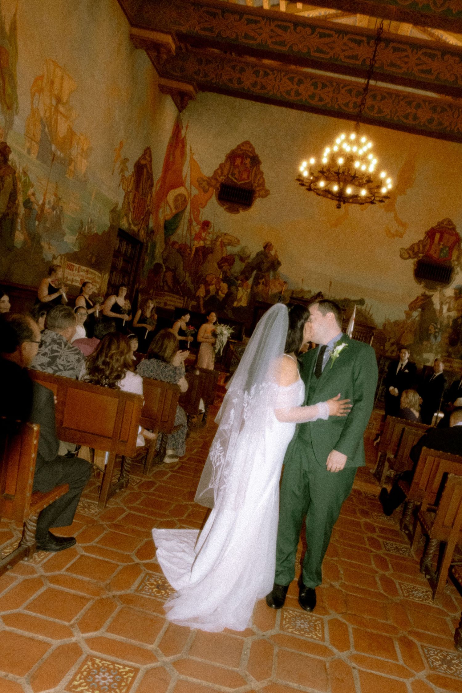 A couple celebrates their wedding moment in an ornate hall with decorative murals and a chandelier overhead.