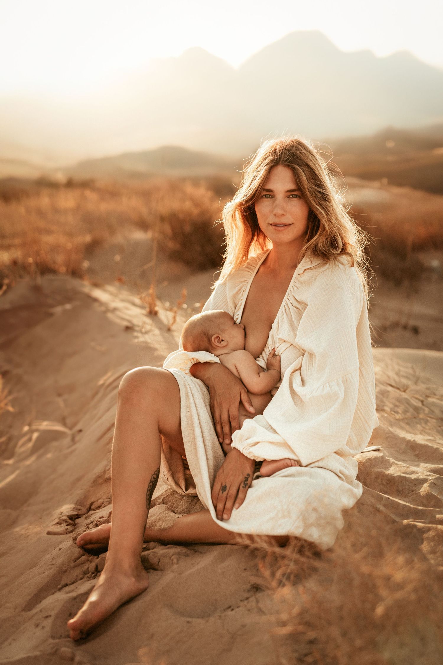 Mother breastfeeding her baby in a white dress while sitting on sand dunes at sunset in golden light. photographer Javea