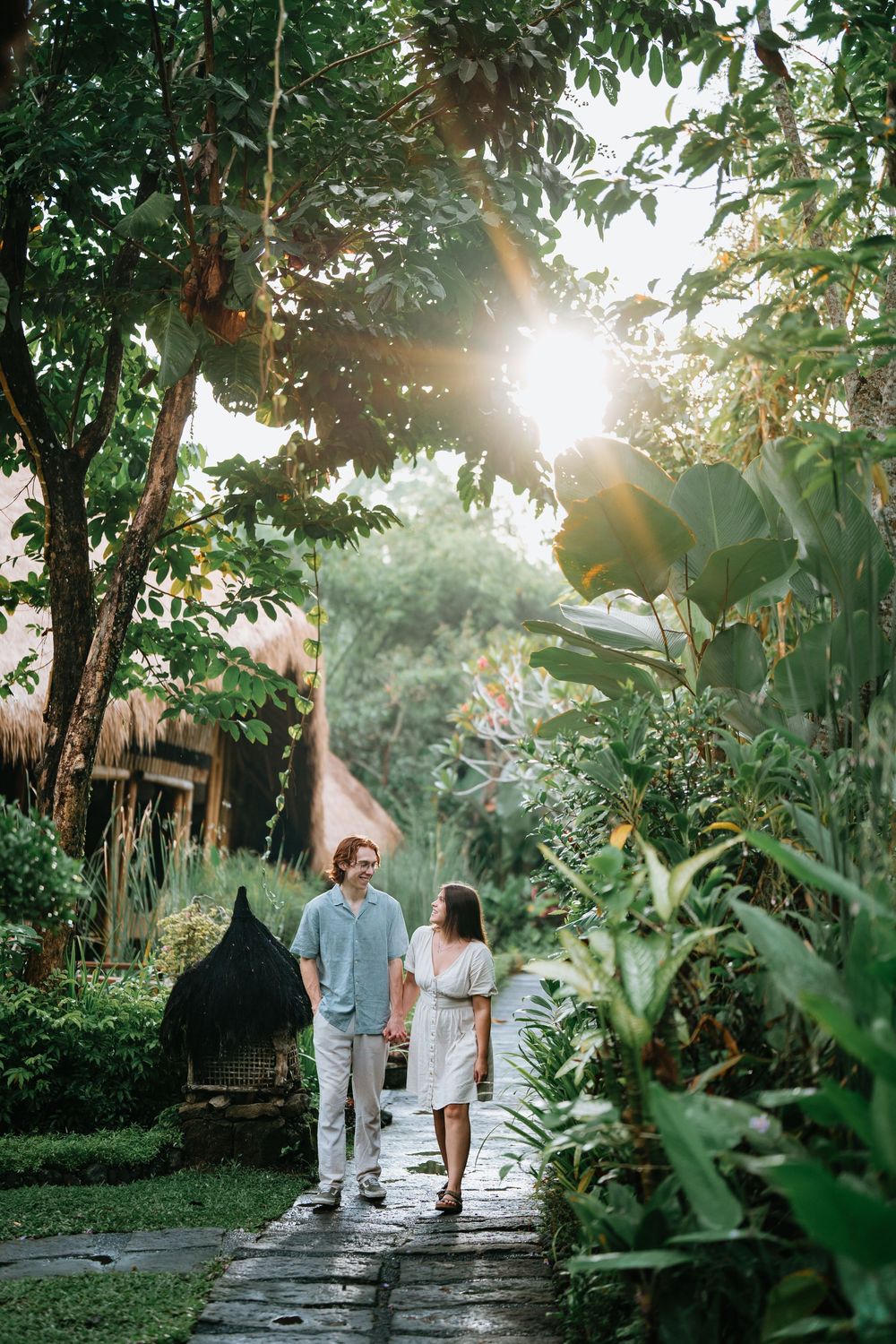 Two people walk hand in hand down a stone pathway surrounded by tropical foliage.