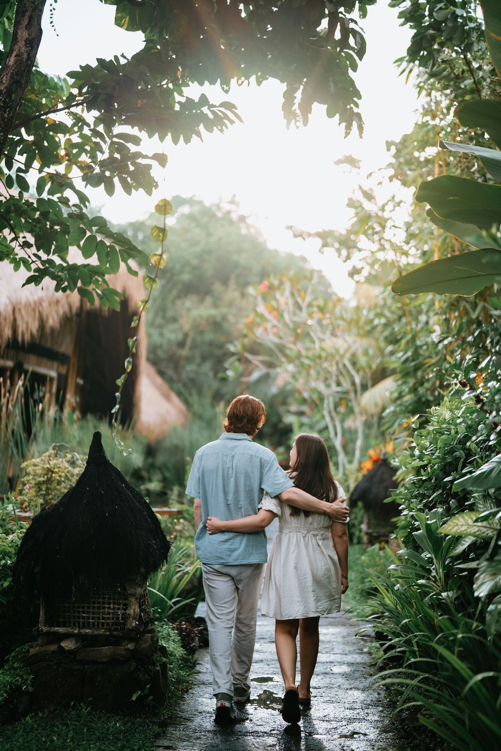 A couple walks hand in hand along a lush tropical garden pathway at sunset.