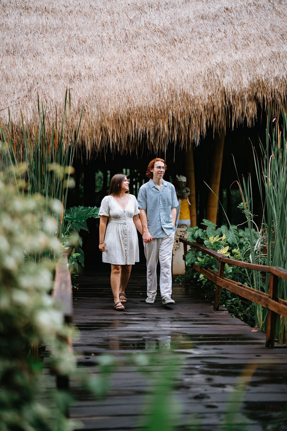 A couple walks along a wooden boardwalk under a thatched roof surrounded by tropical foliage.