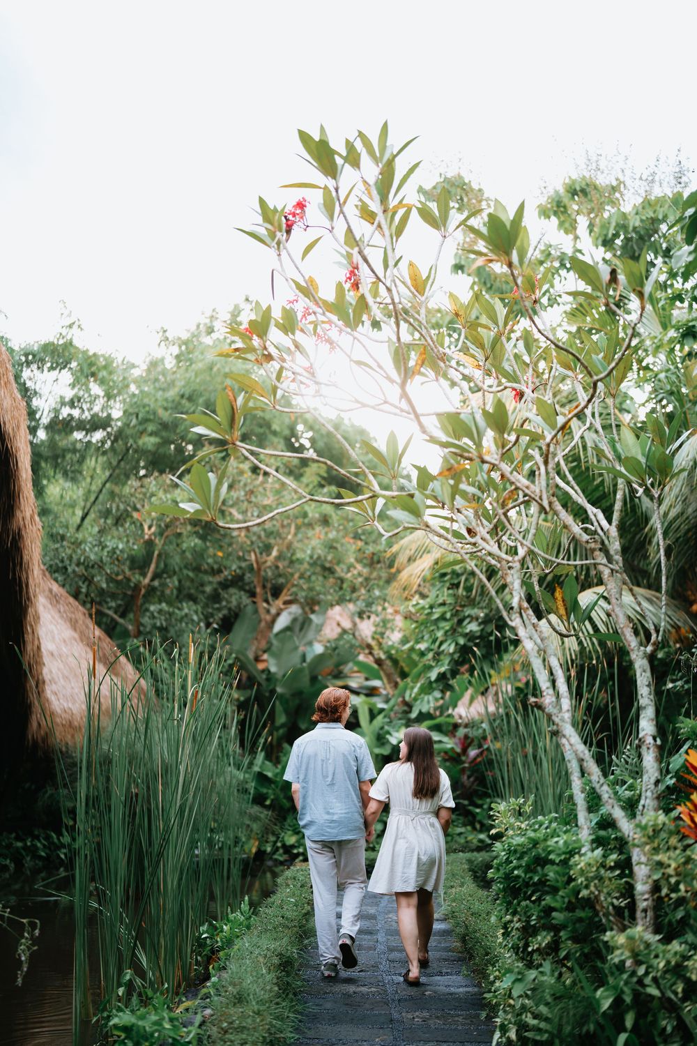 Two people stroll together on a wooden walkway surrounded by tropical plants and bamboo.