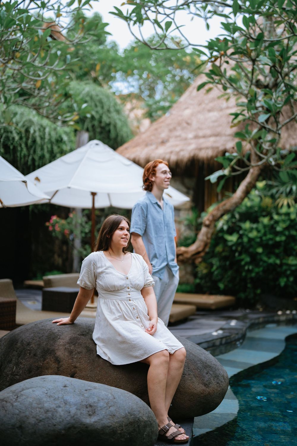 Two people relax by a tranquil pool area with thatched roof bungalows in a tropical resort setting.