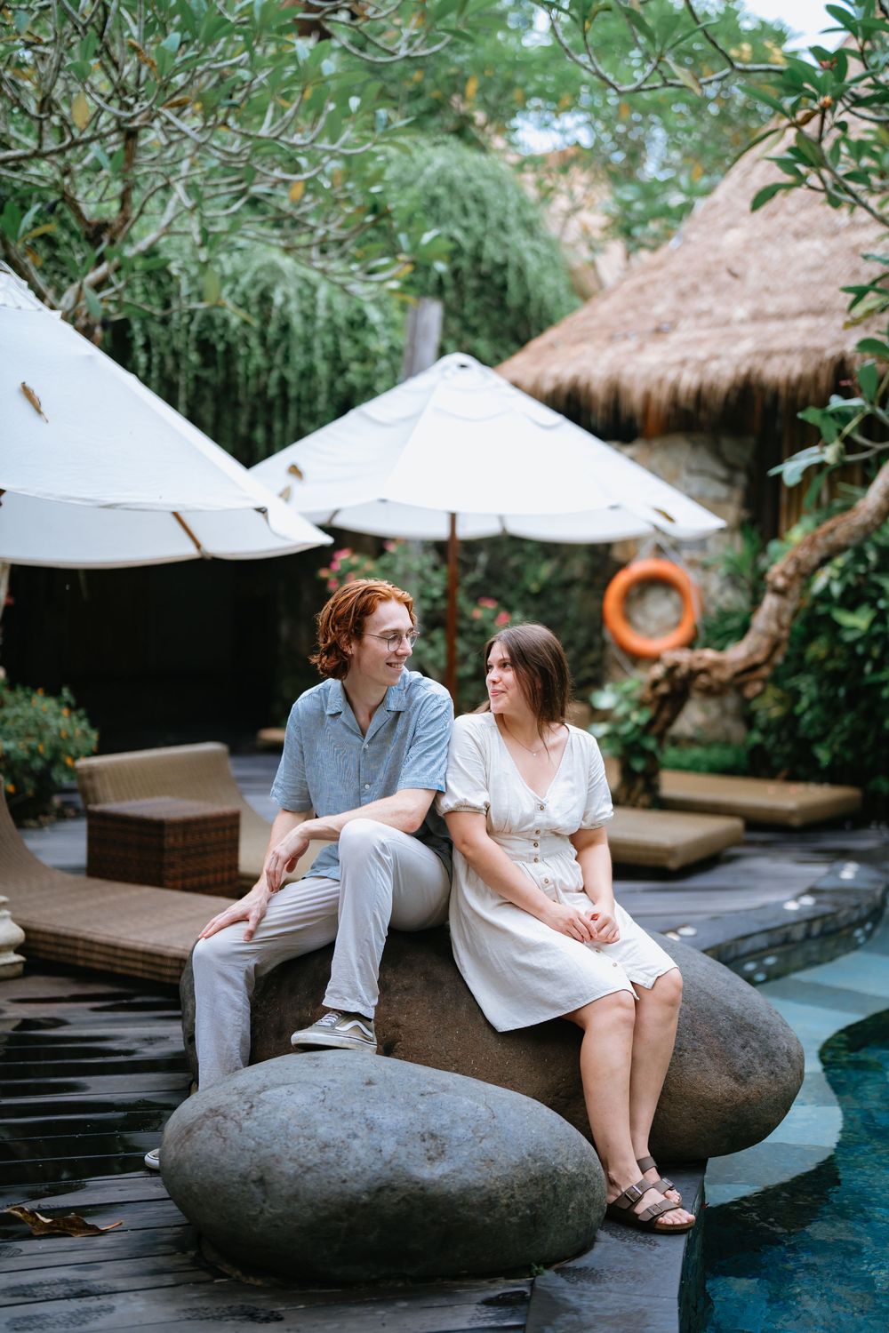 Couple sits on large decorative rocks beside a serene pool at a luxury tropical resort.