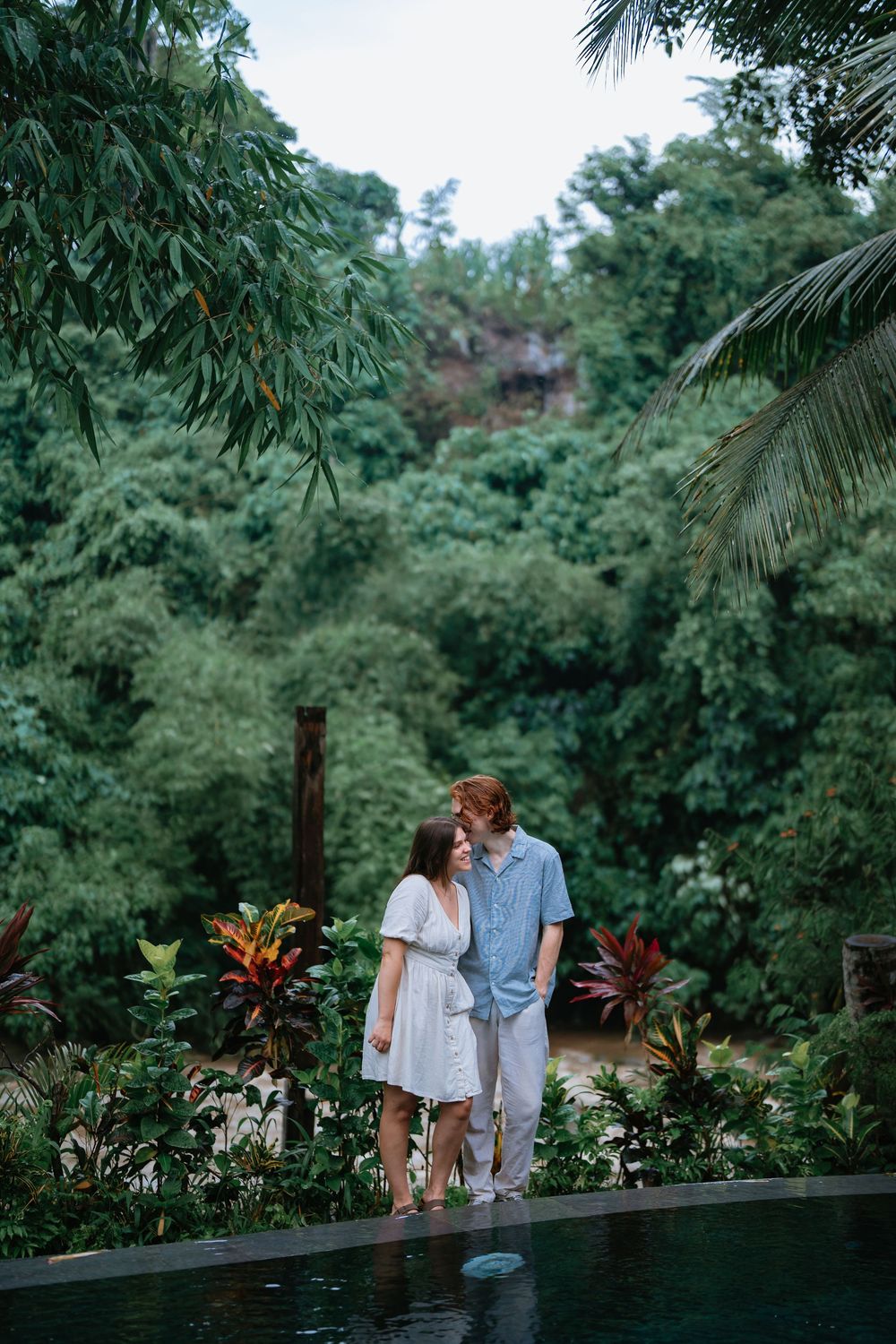 Two people share an intimate moment on a wooden deck surrounded by lush tropical gardens.