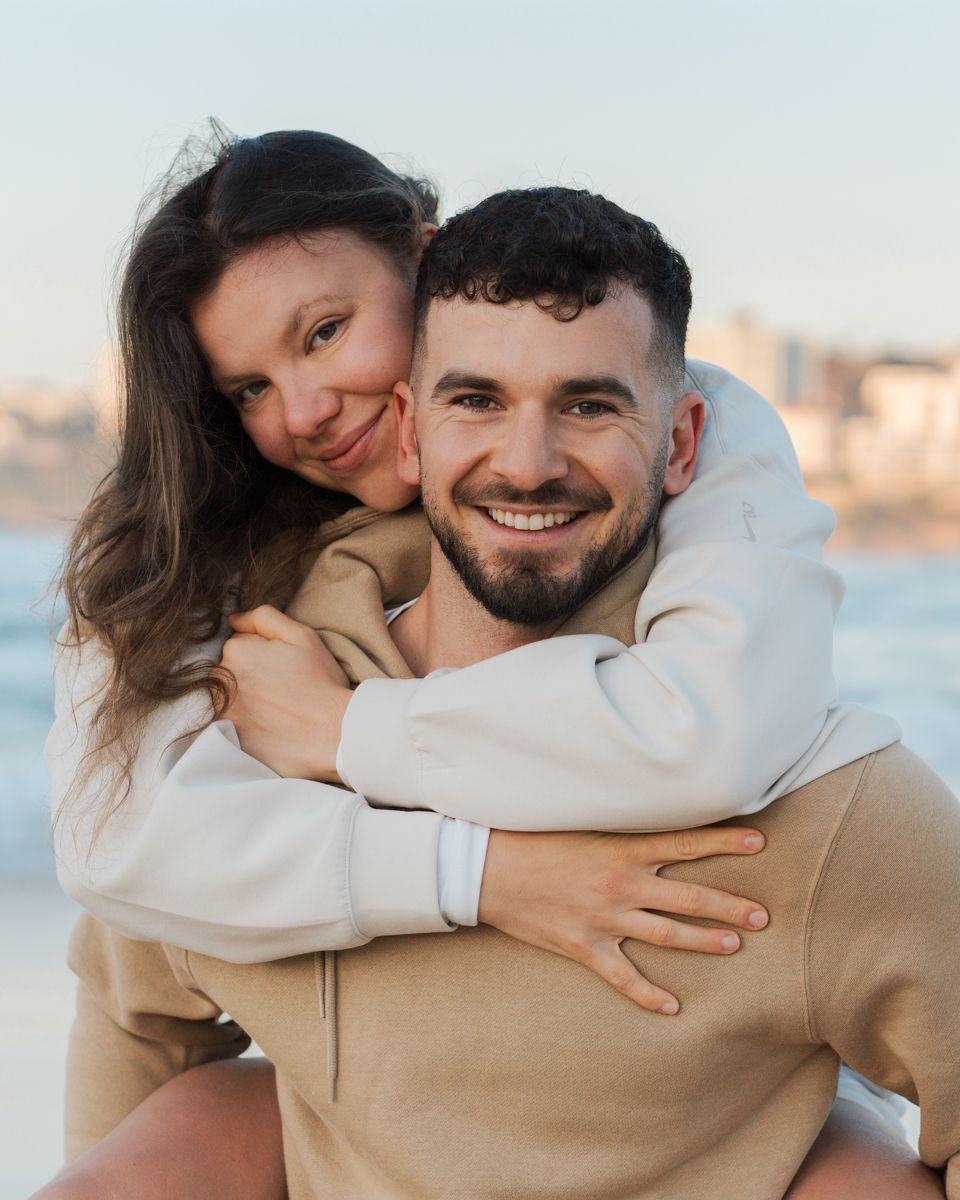 Happy couple hugs outdoors at sunrise, with city skyline and Bondi Beach in the background, wearing matching beige and white sweaters.