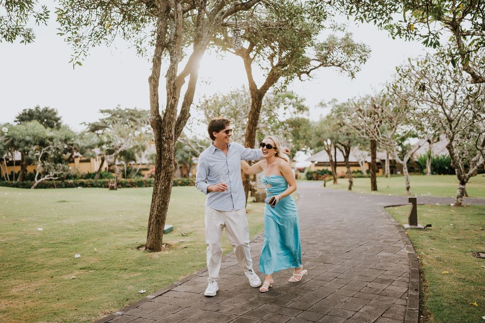 A couple in turquoise and white outfits walk together on a tree-lined path in a park setting.
