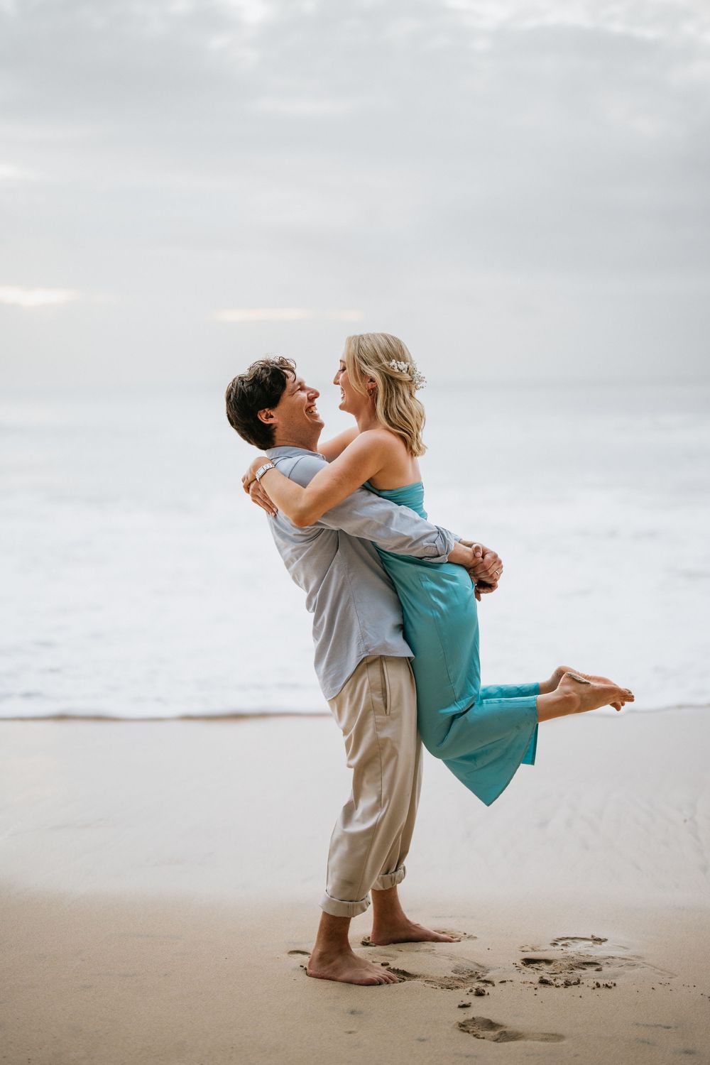 A romantic couple embraces and spins together on an overcast beach.