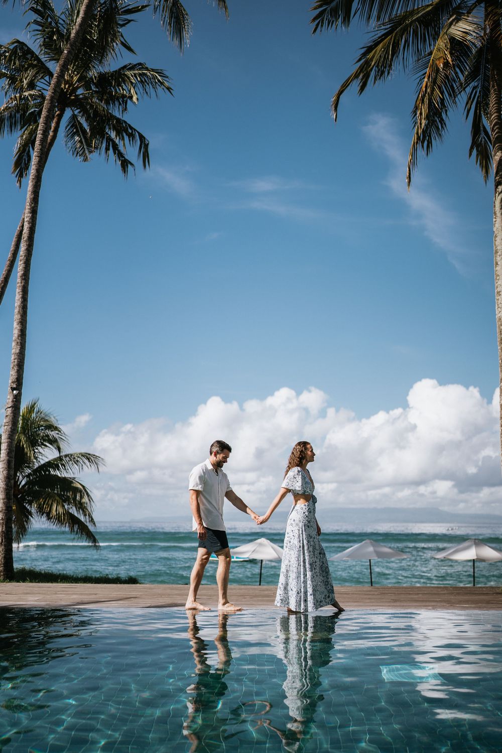 Couple standing near an infinity pool with tropical palm trees and ocean views in the background.