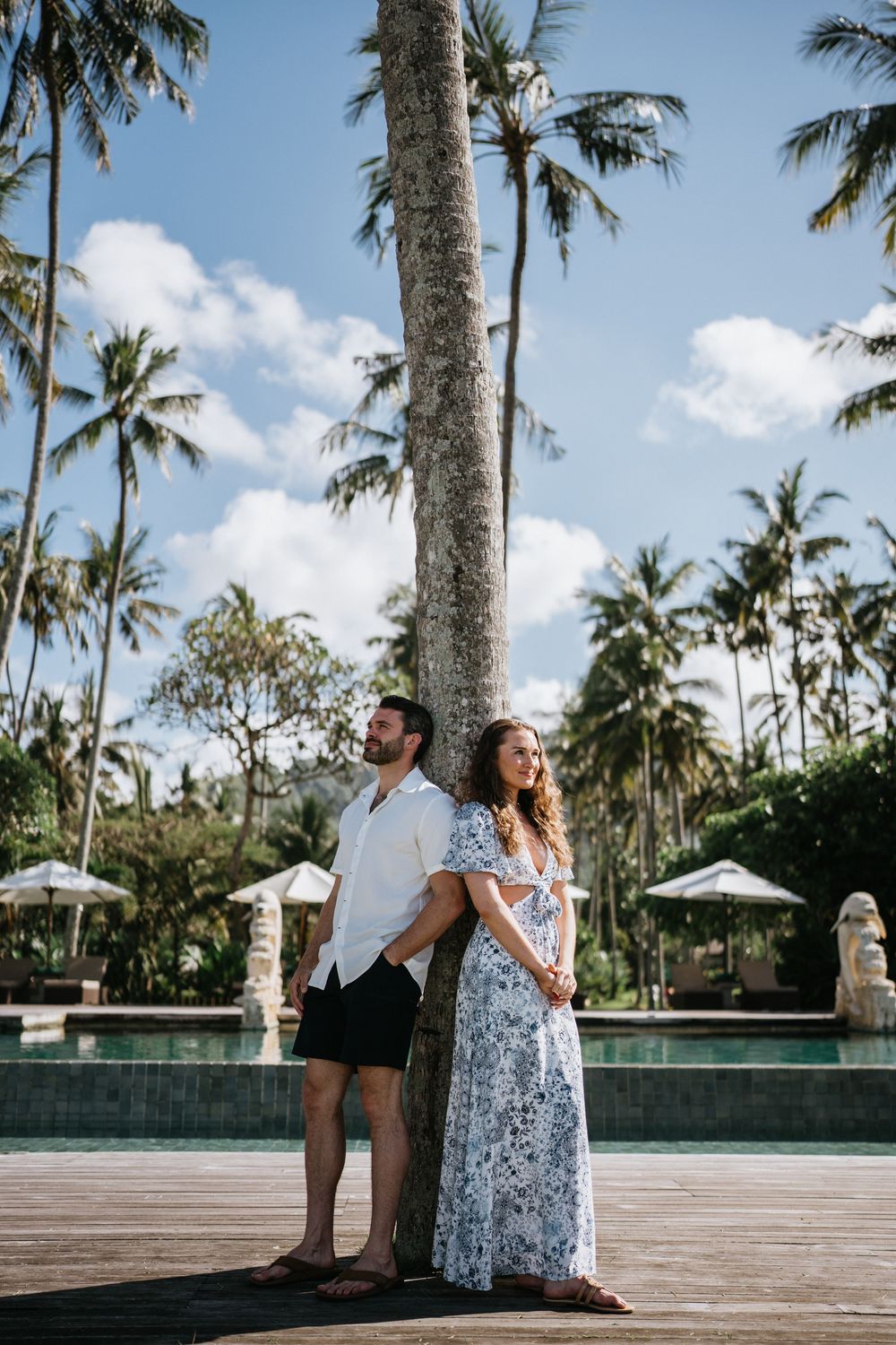 Couple posing on wooden deck near infinity pool with tropical palm grove backdrop.