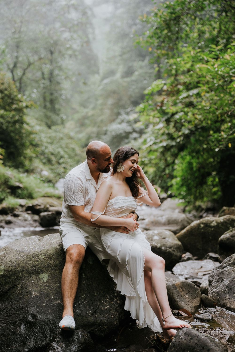 A couple relaxes on large boulders near a forest stream during a romantic photoshoot.