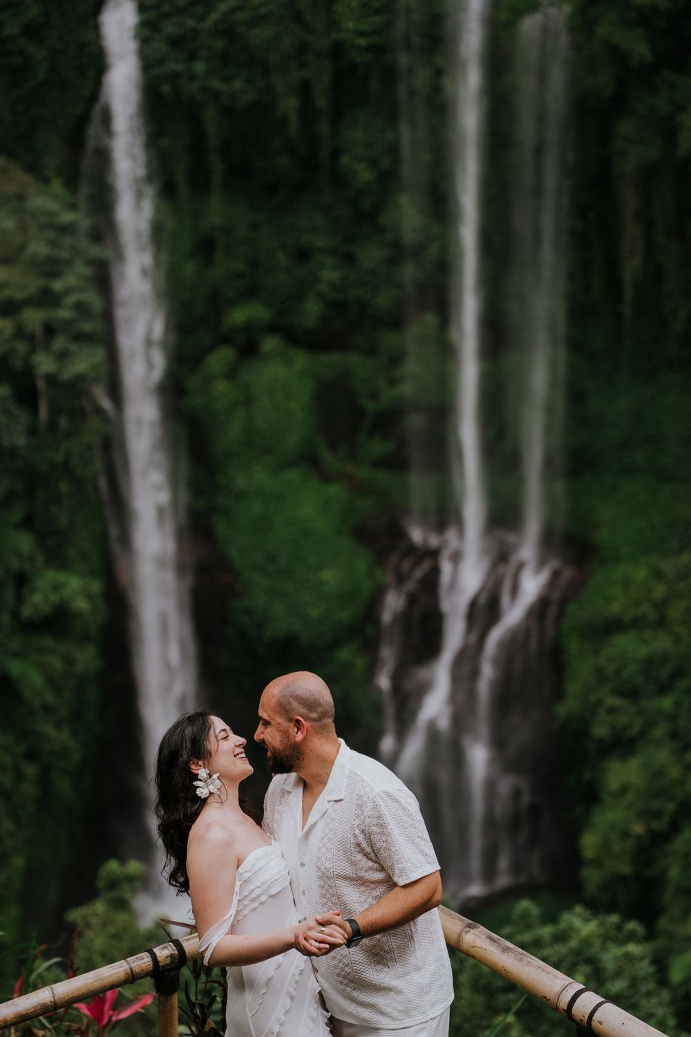 A couple shares a romantic moment in front of a majestic waterfall surrounded by lush greenery.