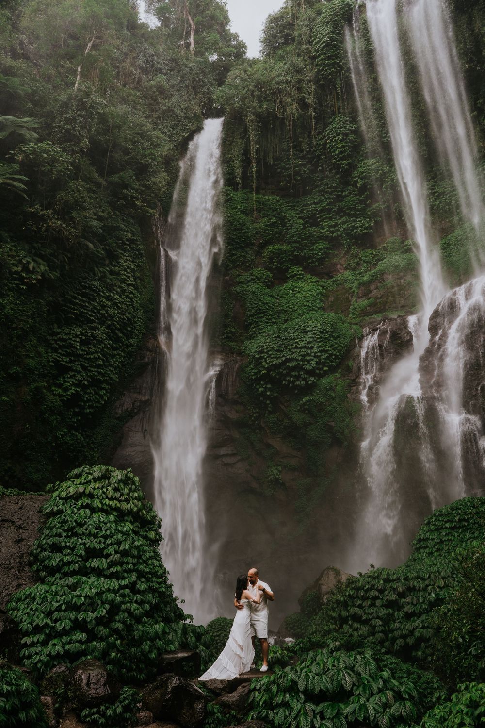 A couple poses romantically in front of a dramatic twin waterfall in a tropical forest.