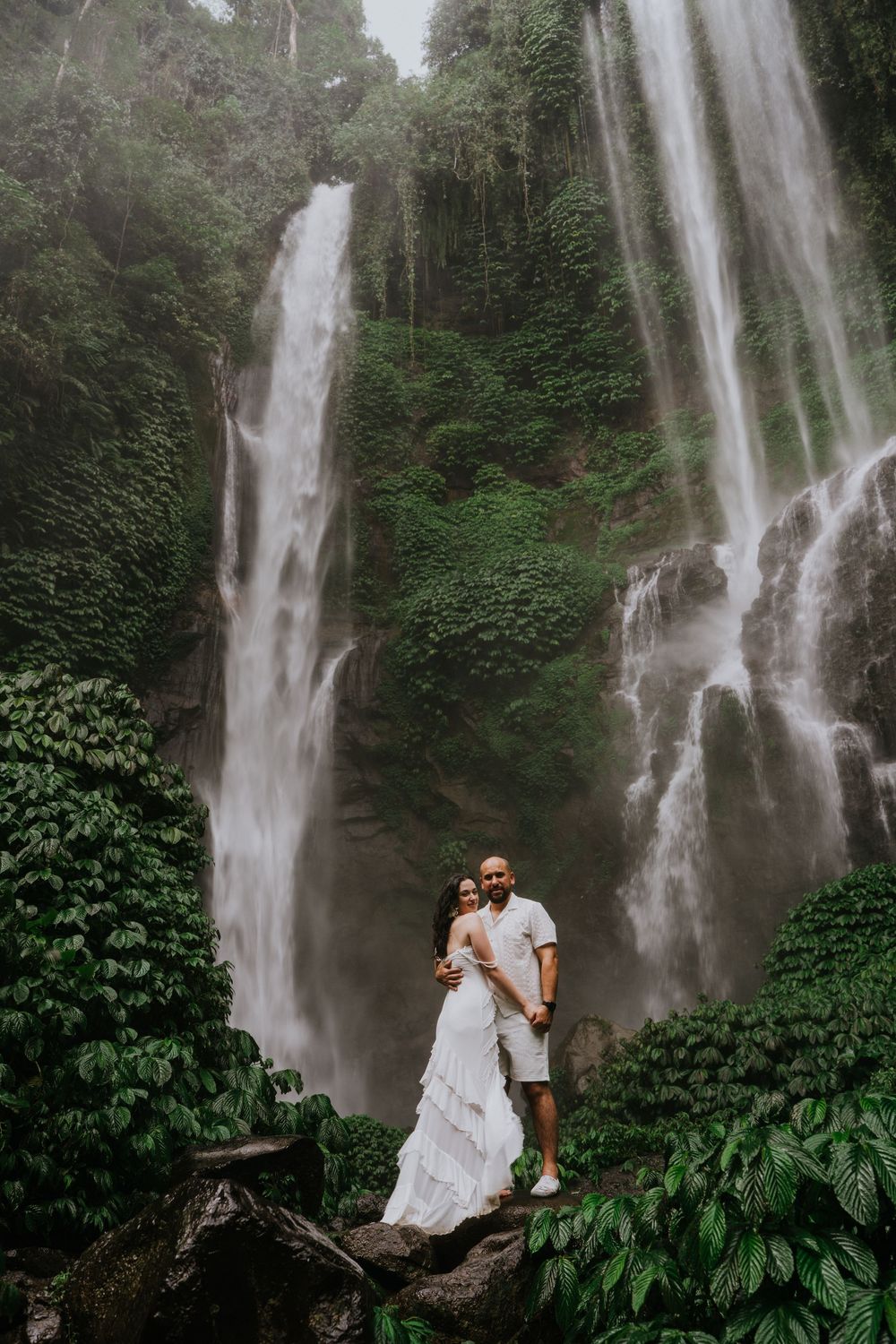 A couple stands on a fallen tree trunk against the backdrop of a magnificent tropical waterfall.
