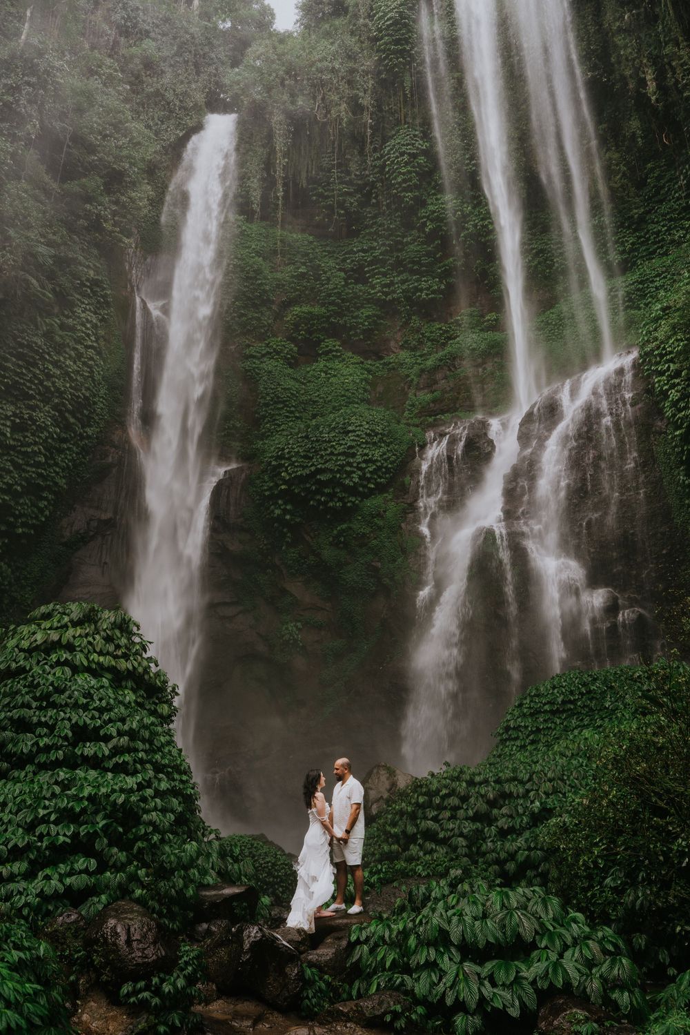 A dramatic waterfall cascades down a lush cliff face surrounded by tropical foliage in a misty forest setting.