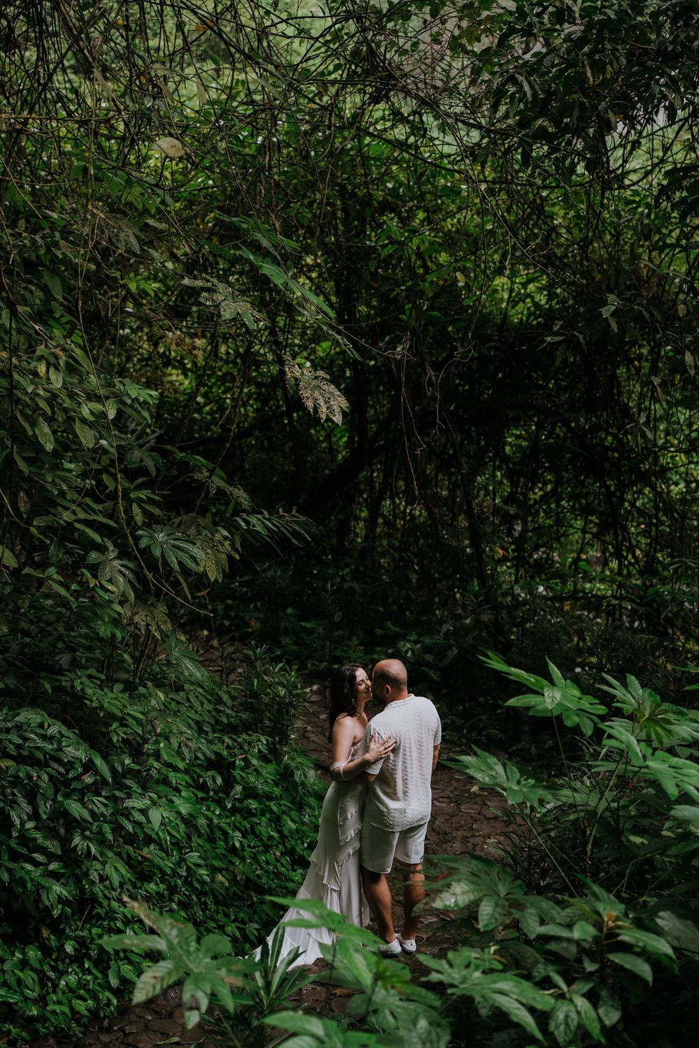 A couple walks along a shaded forest path surrounded by dense tropical foliage.