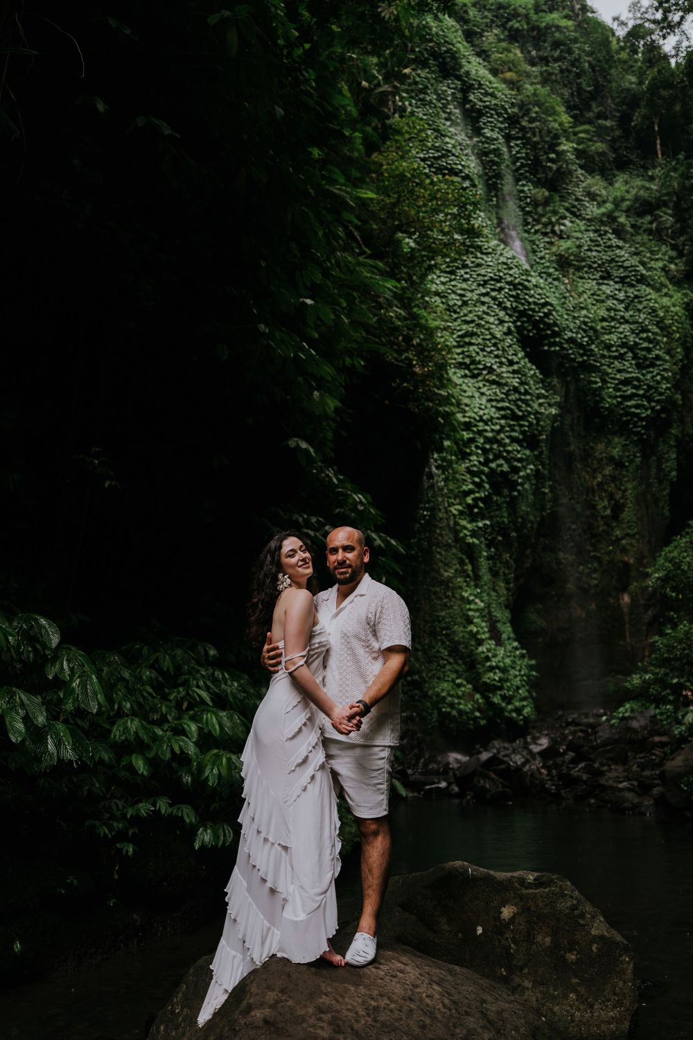 A couple embraces on a rocky outcrop near a scenic waterfall in a tropical setting.