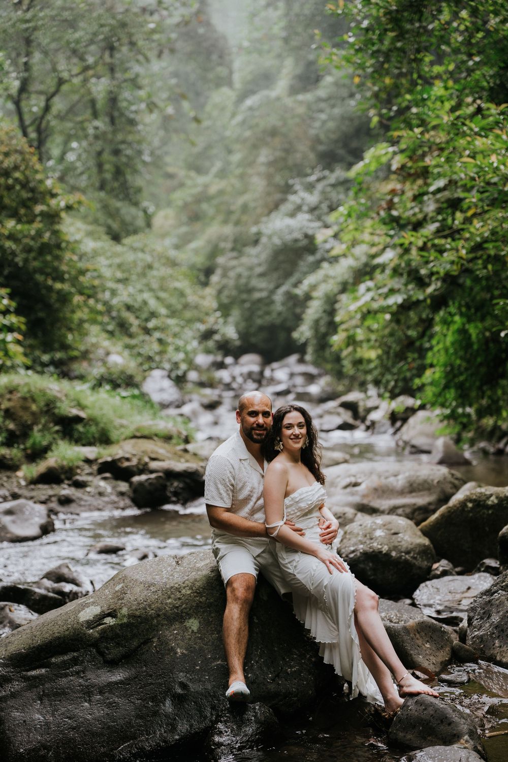 A couple sits together on large rocks by a stream in a misty forest setting.