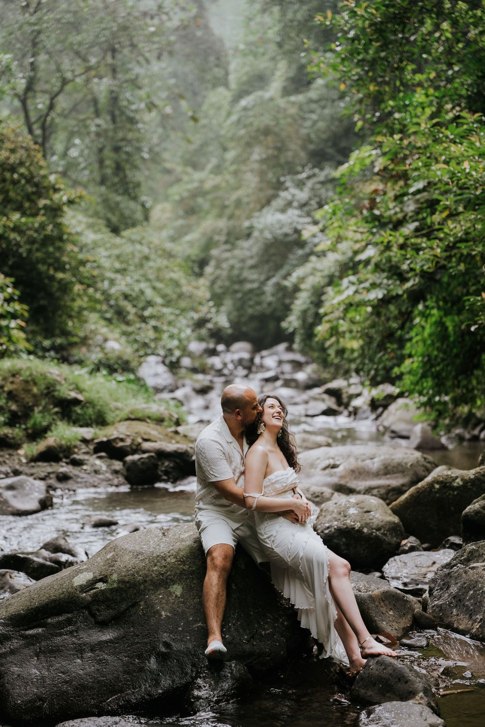 A couple shares an intimate moment while sitting on rocks beside a forest stream.